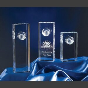 Clear crystal trophy with embedded golf ball, engraved "Presidents Cup First Place," displayed on blue fabric.